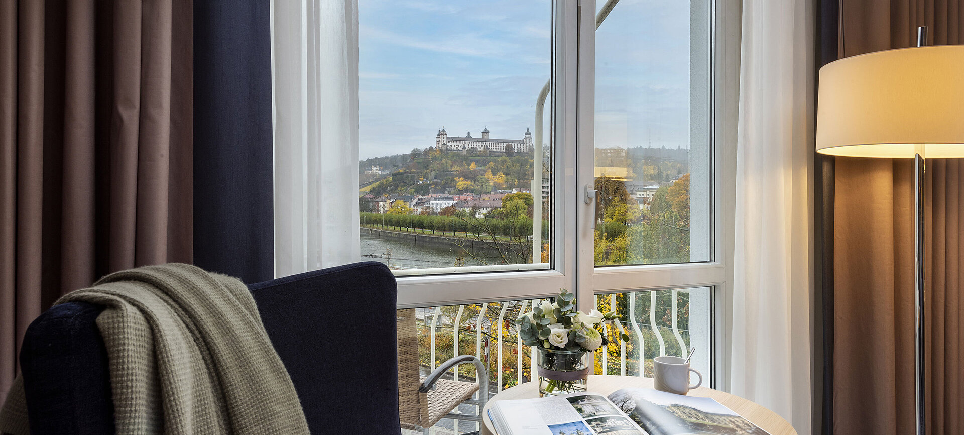 View from a hotel room window overlooking the Main River and Marienberg Fortress on the hill, with a cozy seating area in the foreground